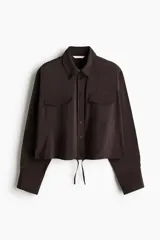 Camisa marrón de manga larga, corte corto y diseño holgado, con cuello clásico, cierre frontal de botones, bolsillos con solapa en el pecho y cordón ajustable en el bajo.