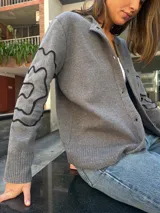 Cárdigan gris de algodón, con cuello redondo, botones frontales y mangas largas con diseño de líneas onduladas en negro.