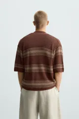 Camisa de punto de manga corta, color marrón rojizo con rayas horizontales en tono beige/verde oliva. Presenta cuello solapa y cierre frontal de botones.