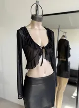 Top negro de mangas largas confeccionado en tejido de red transparente, con volados en el escote y en el ruedo. Se ata en el centro del busto.