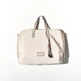 Bolso tipo tote beige con cierre superior, doble asa corta y correa larga desmontable. Incluye charm decorativo con cuentas y flecos.