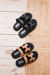 Sandalias planas de cuero sintético, con base negra y tiras anchas en forma de H en color caramelo o negro.