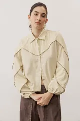 Camisa color beige con cuello camisero, mangas largas abullonadas con puños elásticos y detalles de encaje y costuras en contraste.