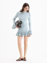 Vestido corto de punto celeste, con silueta ajustada, cuello medio alto y mangas largas. Cuenta con volados plisados en los puños y en el ruedo.