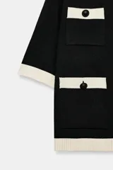 Chaqueta de punto negra de manga corta con detalles en blanco marfil en el cuello, bordes, solapas de los bolsillos y ruedo. Cuenta con cuello redondo, bolsillos delanteros de plastrón y cierre frontal con botones oscuros.
