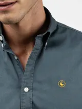 Camisa de hombre color azul, con cuello buttondown, cierre frontal con botones y logo bordado en el pecho. Presenta un acabado vintage logrado mediante el proceso de teñido en prenda.