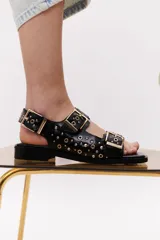 Sandalias negras de cuero con suela plana, dos tiras anchas con hebillas doradas y apliques metálicos.