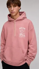 Hoodie color rosado con capucha y cordones ajustables en color crema. Presenta un bolsillo frontal tipo canguro y un estampado gráfico en el pecho con la frase 'Here for the beer'.