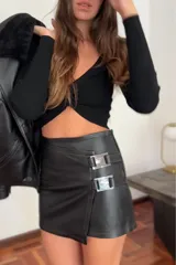 Minifalda negra de cuero sintético, tiro bajo, con corte irregular y detalle de dos hebillas plateadas en el lateral.