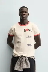 Camiseta de manga corta color crema con cuello redondo y ribetes en color rojo. Presenta un estampado frontal con la palabra "Love" donde la 'o' es un corazón rojo, y varios parches gráficos en tonos grises y negros.