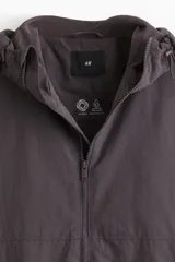Campera rompevientos de nylon repelente al agua, color gris oscuro/marrón topo, con forro de malla. Presenta cierre frontal con protección para el mentón, capucha ajustable con cordón, bolsillos laterales y bolsillo interior con cierre, y elástico revestido en puños y bajo.