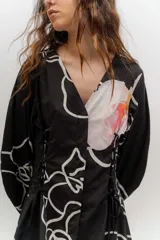 Vestido corto de popelina color negro con estampado de líneas blancas y un diseño floral destacado en el pecho. Cuenta con escote en V, cuello clásico, mangas abombadas y detalles de cordones laterales con efecto corsé.