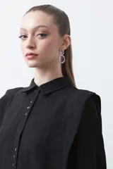 Camisa negra de estilo sastrero con cuello clásico, frente abotonado y mangas largas con puños ajustados. Presenta un diseño con hombreras sutiles que aportan estructura a la prenda.