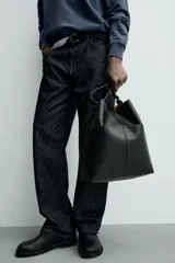 Bolso tipo shopper de cuero sintético negro, con costuras visibles en el frente que dividen el cuerpo en paneles. Presenta un borde superior ondulado y un asa de hombro ajustable.