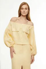 Blusa color amarillo claro, con escote off-shoulder, mangas largas abullonadas y dos bolsillos con solapa en el pecho.
