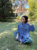 Camisa de denim azul con cuello clásico, mangas largas abullonadas y cierre frontal con botones. Presenta un delicado bordado floral en color rojo a ambos lados del pecho.