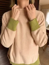 Sweater de punto en color beige con cuello alto y detalles en color verde oliva en los puños y el ruedo. Presenta un diseño holgado con parche de marca en el lateral.