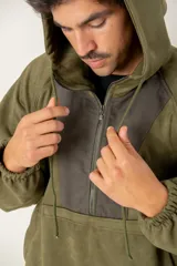 Canguro con capucha de estilo urbano en color verde oliva, confeccionado en tejido suave. Presenta un panel frontal en contraste de color gris oscuro con cierre de cremallera parcial, mangas largas con puños elásticos y bolsillos laterales integrados.