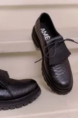 Mocasín de cuero vacuno negro con textura tipo reptil, suela track de goma, cordones finos y solapa con flecos sobre el empeine.