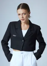 Saco sastrero corto a la cintura, color negro, con cierre frontal de un botón grabado, solapas clásicas, bolsillo ojal en el delantero, chapa metálica con logo en el pecho y bordado decorativo en la manga.