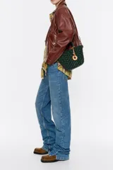 Bolso tipo cesta de estructura rígida y diseño trenzado en color negro. Cuenta con dos asas superiores cortas y una correa larga ajustable en color marrón. Incluye un dije circular decorativo con grabado en tono madera.