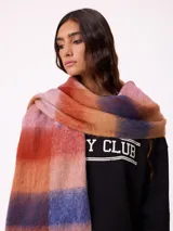 Bufanda de tejido suave con diseño de cuadros grandes en tonos naranja, rojo, violeta y lila. Presenta flecos en los extremos y una etiqueta pequeña con el logo de la marca Rusty.