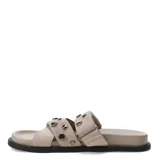 Sandalias planas de mujer de estilo urbano, confeccionadas en material sintético símil gamuza de color beige. Presentan un diseño destalonado con dos tiras anchas en el empeine decoradas con tachas metálicas oscuras. Cuentan con una base plana con plantilla anatómica para mayor comodidad.