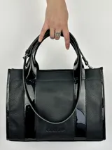 Bolso tote negro de cuero con detalles de charol, doble asa corta y correa bandolera de tela con letras.