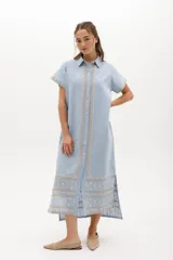 Vestido camisero de lino color lila con bordados rojos en contraste. Presenta un diseño de largo midi con cuello camisero, cierre frontal de botones y mangas cortas. Los detalles bordados recorren la botonadura, los puños y el ruedo de la prenda.