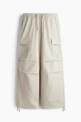Pantalón cargo de algodón color beige con corte holgado. Presenta cintura elástica con cordón ajustable, múltiples bolsillos con solapa y botones a presión en las piernas y parte trasera, pliegues en las rodillas y cordones de ajuste en los bajos.