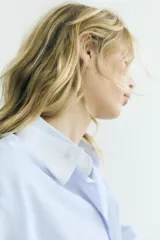 Camisa oversize de algodón con estampado de rayas finas, cuello solapa, escote en pico y manga larga con puños abotonados. Presenta un diseño con pliegues laterales y cierre frontal con botones.