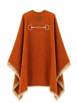Ruana de algodón color naranja con diseño de tejido en espiga y detalle de estribo bordado en la parte superior. Presenta terminaciones con flecos en tono beige en todo el contorno.