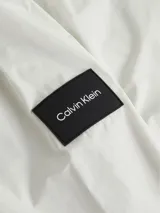Campera blanca de poliéster con capucha y cierre frontal con cremallera. Tiene cuello alto, puños y ruedo elásticos, y un parche de goma con el logo de Calvin Klein en la manga izquierda.