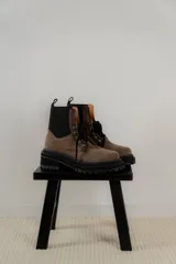 Borcego de gamuza color chocolate con paneles laterales elásticos negros y suela track dentada. Presenta sistema de cordones con ganchos metálicos y tirador en la parte trasera.