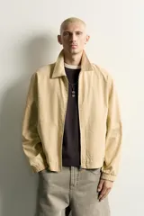 Cazadora regular fit de color beige, confeccionada en mezcla de algodón y lyocell. Cuenta con cuello camisero, cierre frontal de cremallera, bolsillos de plastrón en la cadera, mangas largas con puños elásticos y bajo con ajuste elástico.