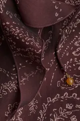 Vestido largo de manga globo, color marrón oscuro, con estampado paisley en tonos claros. Presenta cuello alto con volados y escote en V con lazos ajustables, y puños con volados.