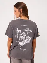 Remera gris con efecto acid wash, corte loose y estampado gráfico trasero con ilustraciones de muebles de playa y textos en blanco.