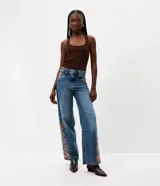Pantalón wide leg de jean azul con bordado floral aplicado en los laterales. Posee cintura mediana con trabillas, cierre frontal con botón y cremallera, y bolsillos funcionales.