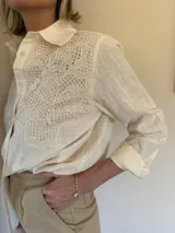 Camisa de manga larga en color beige, con cuello clásico y cierre frontal de botones. Presenta un delicado aplique de encaje tipo crunie en la parte superior delantera, aportando textura y un detalle artesanal al diseño.