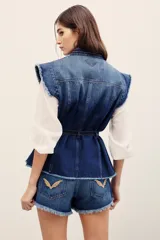 Chaleco de denim azul de corte estructurado y sin mangas, con cuello mao y cierre frontal con botones. Presenta bolsillos frontales con solapa y detalles deshilachados en las sisas y el ruedo.