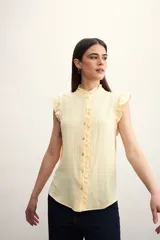 Camisa color amarillo pálido con textura arrugada, cuello mao con volados, cierre frontal con botones y mangas cortas con volados.