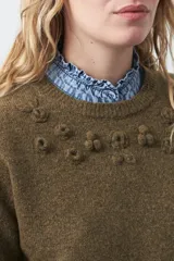 Sweater de tejido suave color tostado, con cuello redondo y detalle de flores bordadas en relieve en la parte superior del pecho. Presenta un corte relajado y mangas largas con puños ajustados.