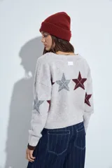 Cardigan corto tejido en color gris, con escote en V y cierre frontal de botones. Presenta un diseño estampado con estrellas y líneas geométricas en tonos oscuros y rojizos, con mangas largas y puños acanalados.