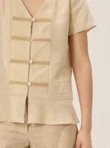 Camisa de manga corta color beige con estampado a cuadros, cuello en V y detalles de galones decorativos estilo oriental en la parte frontal.