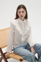 Camisa de algodón blanca con cuello camisero, cierre frontal de botones y volados bordados en el frente y hombros. Presenta mangas largas con puños acampanados y detalles bordados en el ruedo.
