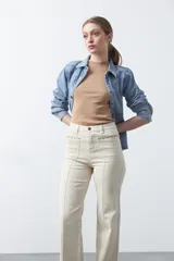 Pantalón de fit recto color beige, confeccionado en cotton spandex. Presenta costuras visibles y bordados decorativos en tono dorado/marrón claro. El ruedo del pantalón tiene un borde festoneado.