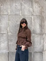 Blusa marrón de manga larga con diseño fruncido en el pecho y cuello alto con volados, estilo romántico y mangas abullonadas con puños ajustados.