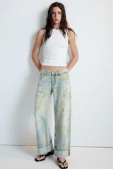 Jeans de tiro bajo confeccionados en denim, con diseño holgado y estampado integral de rosas en tonos amarillos y verdes sobre fondo celeste claro. Cuenta con cinco bolsillos, trabillas para cinto y botamangas con vuelta.