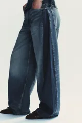 Jeans de tiro alto, color azul medio lavado, con un corte extremadamente ancho y voluminoso (balloon/oversize). Presenta costuras torcidas y paneles de denim de tono ligeramente diferente en los laterales, creando un efecto visual dramático.