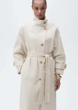 Abrigo largo de diseño oversize en color beige, confeccionado en tejido de algodón con elastano. Presenta cuello alto abotonado, cierre frontal con botones, mangas largas con trabillas en los puños y cinturón tipo batín para ajustar a la cintura. Incluye dos bolsillos laterales.